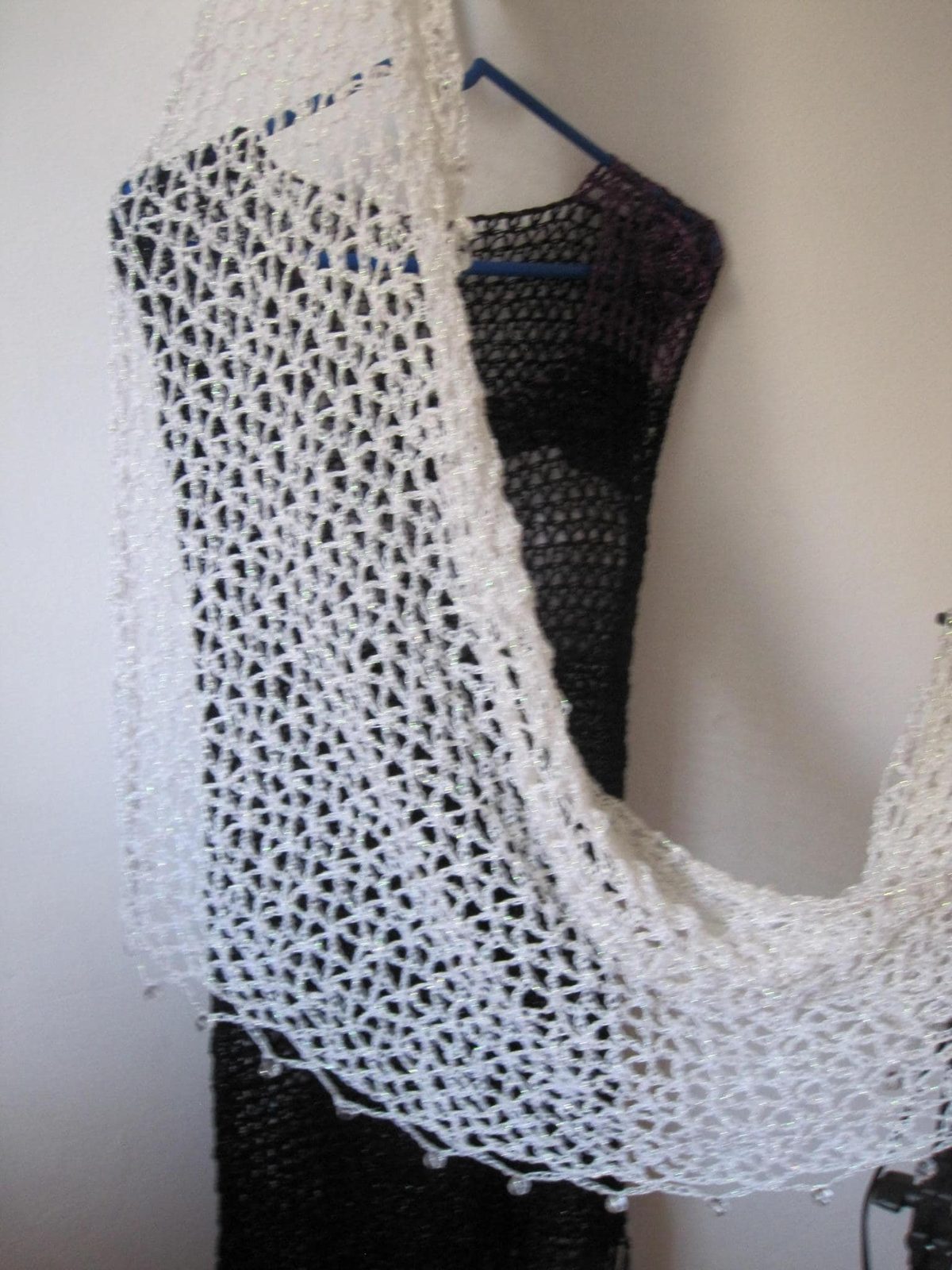 Cream crochet shawl draped over a black dress on a hanger, side view of the airy openwork wrap.